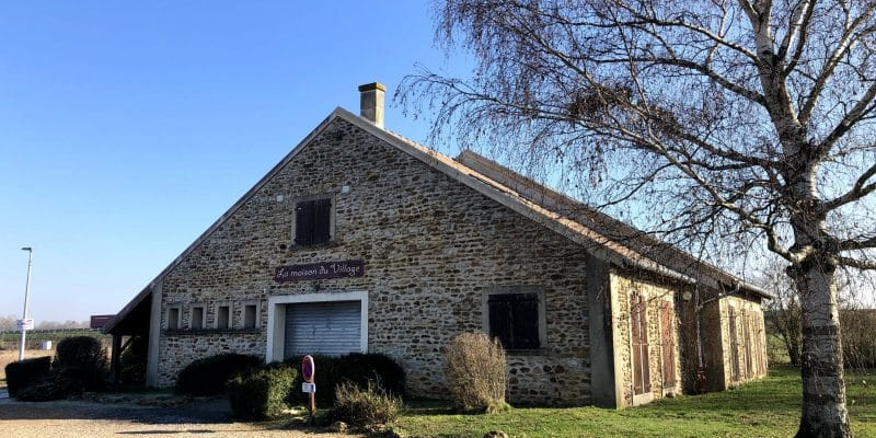Bâtiment en pierre avec un panneau, un arbre nu, des buissons et une allée en gravier.