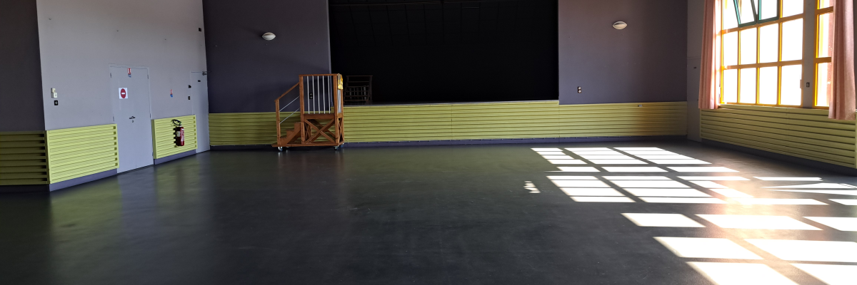 Salle avec sol noir, murs partiellement jaunes, fenêtres, porte, escalier et projecteurs suspendus.