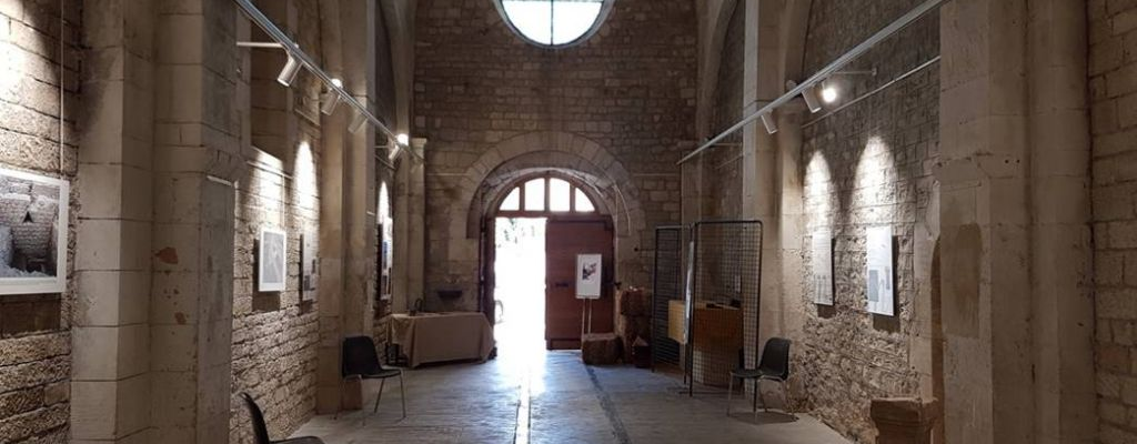 Salle avec murs de pierre, vitraux, lumières suspendues, œuvres encadrées, chaises, canapé et tables.