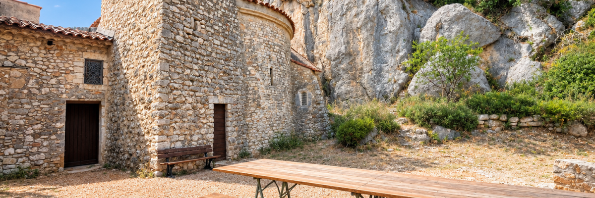 Ancienne église en pierre avec bancs et tables en bois à l'extérieur, rochers et végétation.