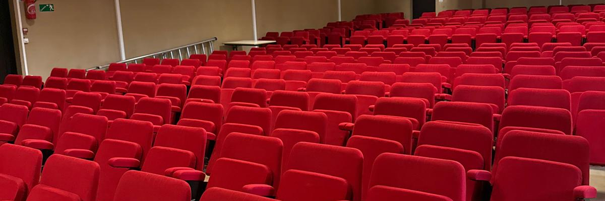 Salle avec rangées de chaises rouges, table à l'avant, écran et porte.