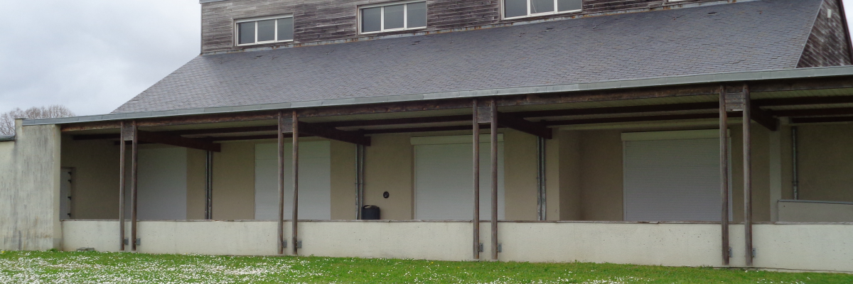 Bâtiment avec toiture en pente, murs blancs, avant-toit soutenu par des piliers. Pelouse avec fleurs sauvages.