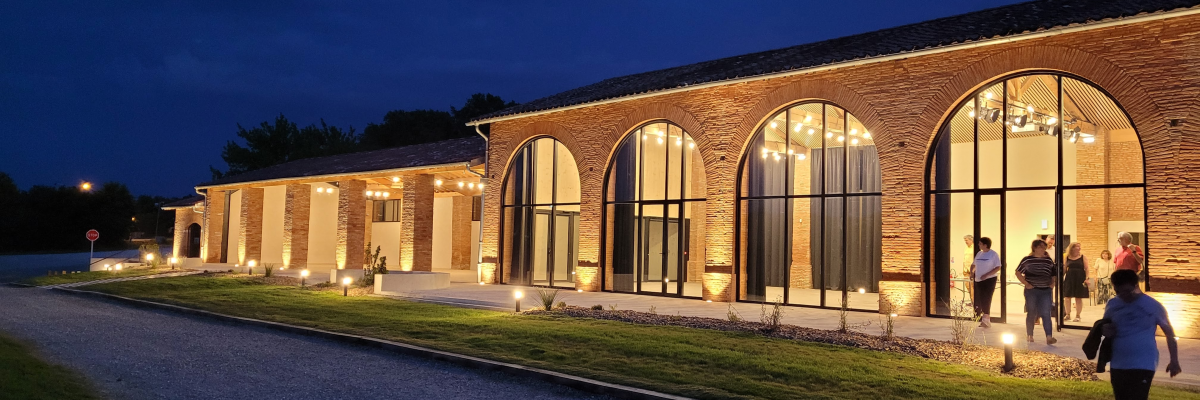 Bâtiment en brique avec grandes fenêtres éclairées, allée et pelouse.