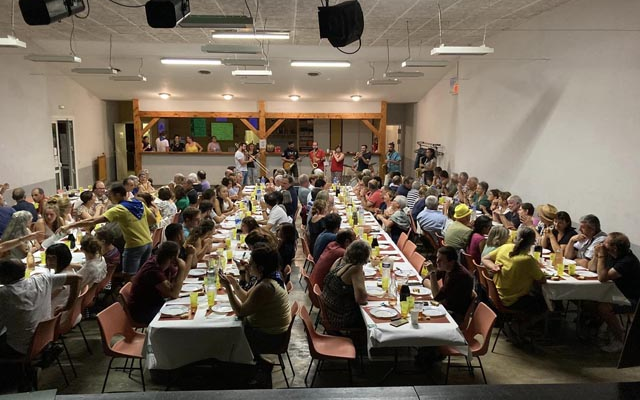 Salle avec tables, chaises, projecteurs suspendus, personnes assises et mangeant.