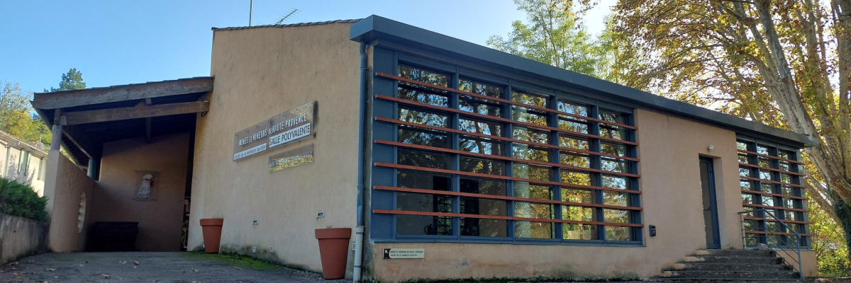 Bâtiment avec grandes fenêtres, auvent, pots de fleurs et un panneau.