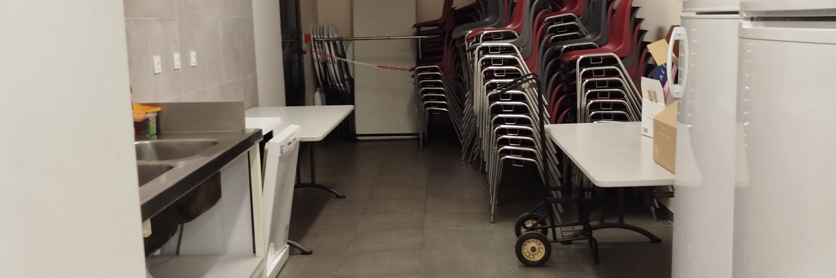 Pièce avec miroir, lavabo, tables pliantes, chaises, réfrigérateur, chariot et étagères.