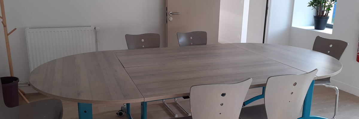 Salle de réunion avec table ovale, chaises, porte-manteau, radiateur et plante.