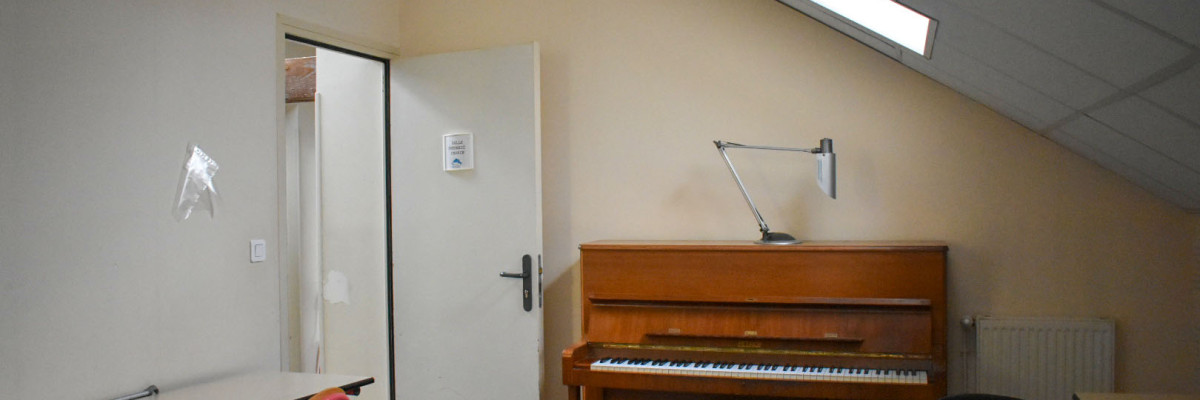 Salle avec piano, tabouret, lampe, table, chaise, radiateur et porte.