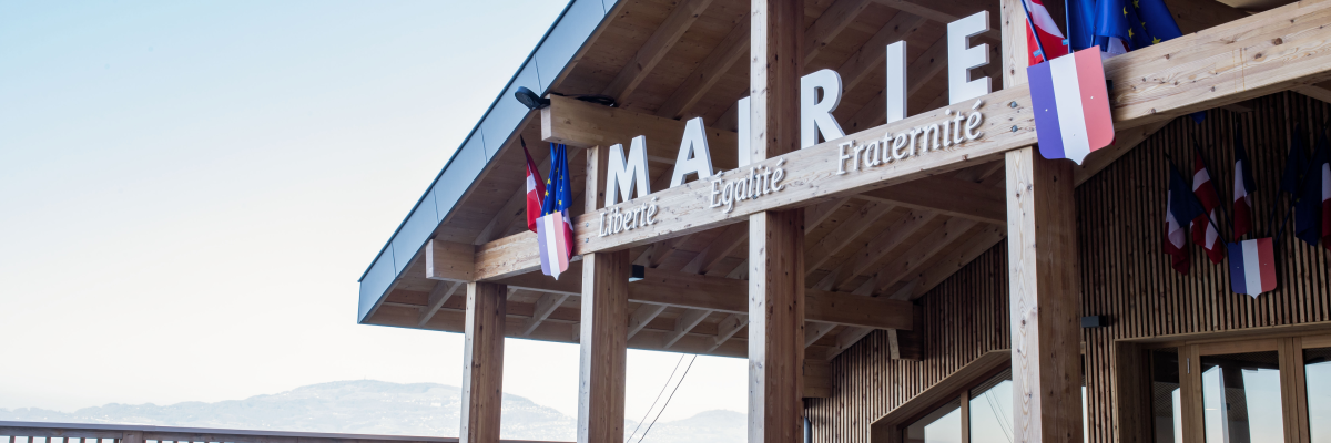Bâtiment en bois avec enseigne "Mairie Liberté Egalité Fraternité", drapeaux, balcon et vue sur montagnes.