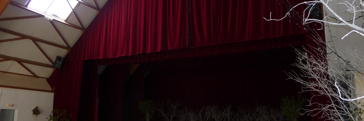 Salle avec scène, rideau, arbre décoratif, vitrail, porte.