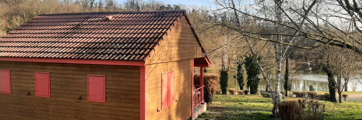 Petite maison en bois avec toiture en tuiles, portes rouges, située dans un jardin avec arbres et autre habitation au loin.