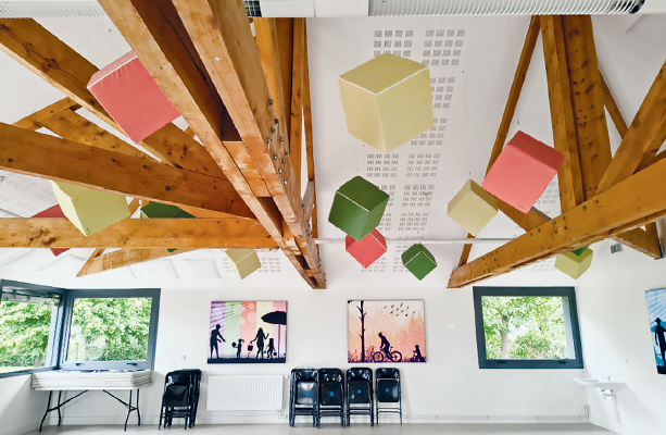 Salle avec poutres apparentes, cubes colorés suspendus, chaises, table, tableaux et fenêtres.
