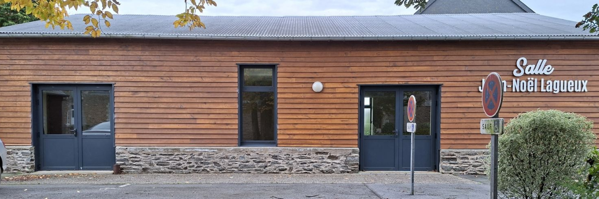 Bâtiment en bois avec toit métallique, trois portes bleues, deux panneaux de signalisation et une voiture à gauche.