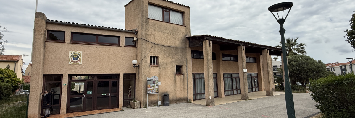 Bâtiment municipal avec entrée, drapeaux, lampadaire et végétation.