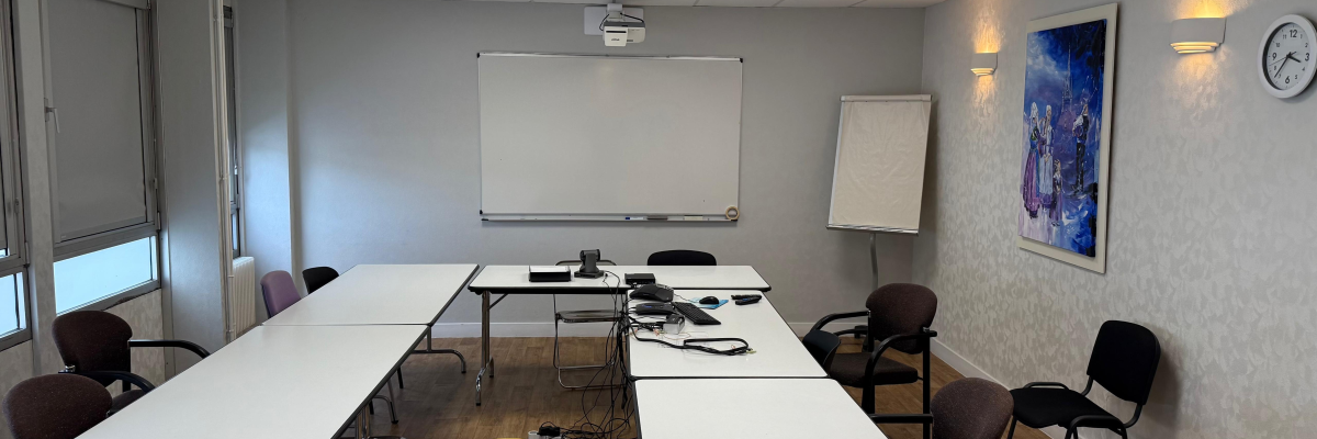 Salle avec tables, chaises, vidéoprojecteur, tableau blanc, paperboard et horloge.