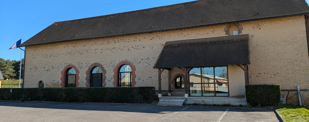 Bâtiment en briques avec trois arches vitrées, toit en pente, entrée avec auvent, parking et haie.