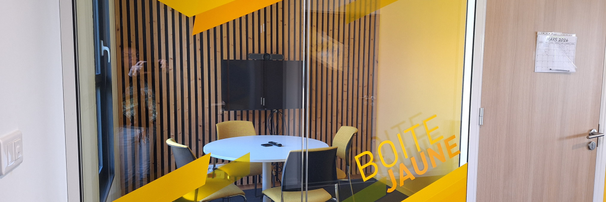 Salle de réunion avec table ronde, chaises, écran. Porte en verre avec motifs jaunes.