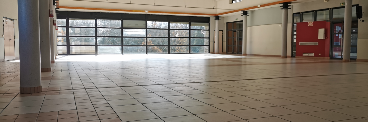 Grand espace avec sol carrelé, grandes fenêtres, piliers, plafond en arc avec poutres en bois, extincteur et distributeur automatique rouge.