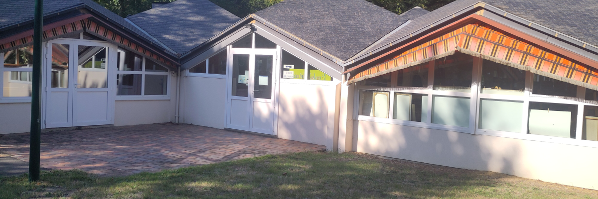 Bâtiment avec toits pentus, murs blancs, fenêtres et portes vitrées. Pelouse et arbres en arrière-plan.