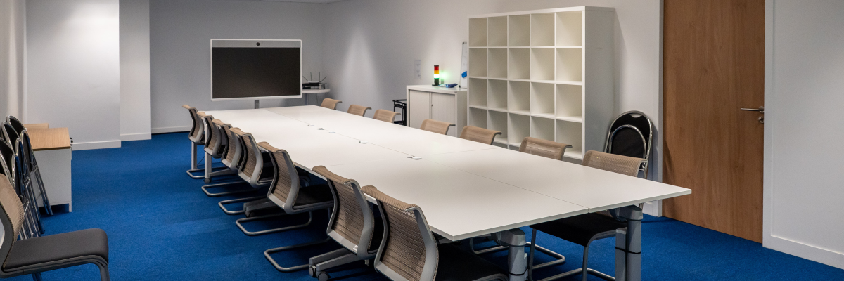 Salle de réunion avec table, chaises, écran, étagère et porte.