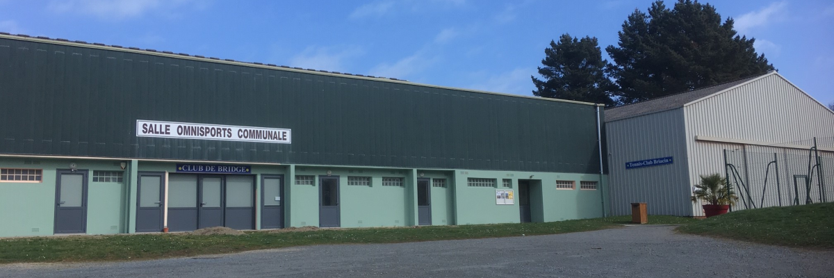 Bâtiment vert avec plusieurs portes, situé dans une zone pavée, sous un ciel bleu avec des nuages.