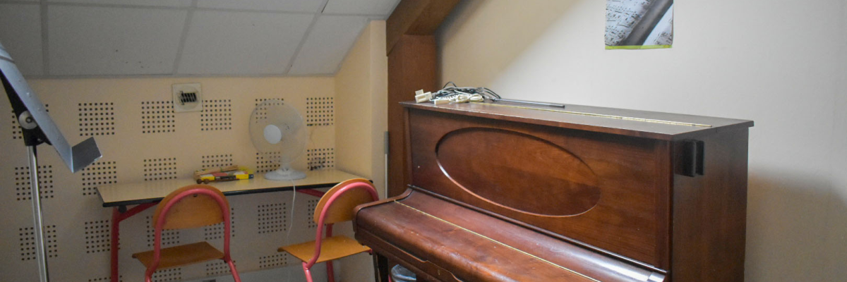 Piano avec tabouret, bureau, chaises, ventilateur, tableau.