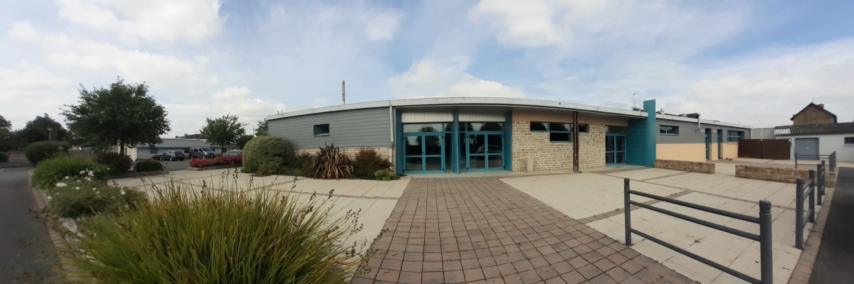 Bâtiment avec entrée, chemin de briques, plantes, voiture au loin, ciel partiellement nuageux.