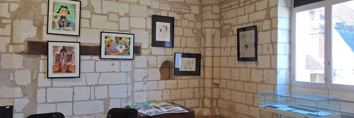 Salle d'exposition avec murs en pierre, tableaux, vitrine et table avec brochures.