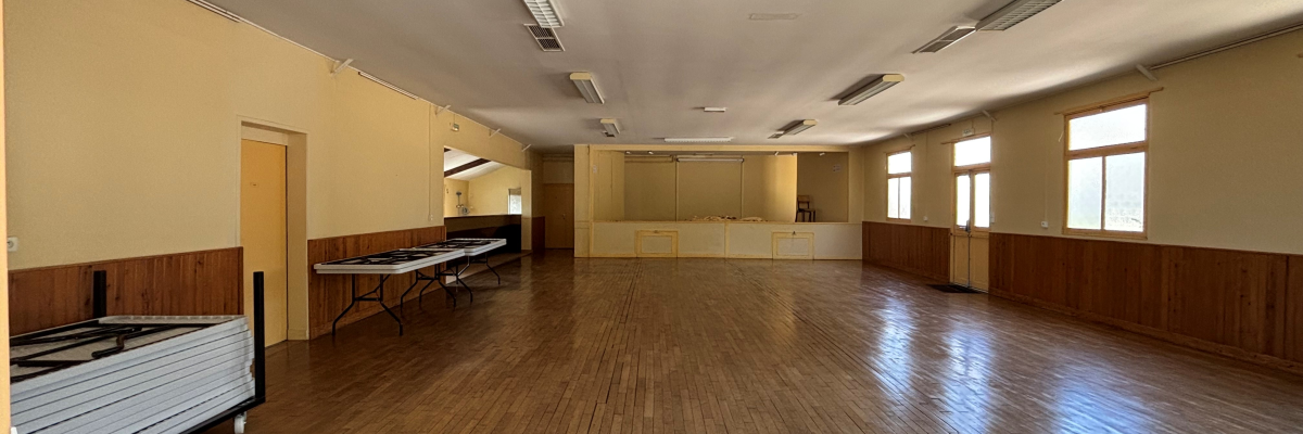 Salle vide avec parquet, tables, chaises, et fenêtres.