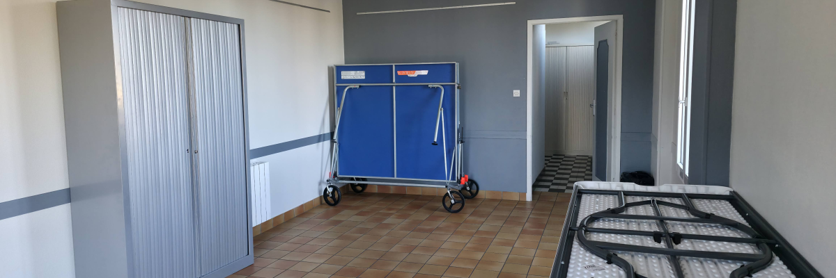 Pièce avec armoire, chariot et sommier. Sol carrelé, murs gris.