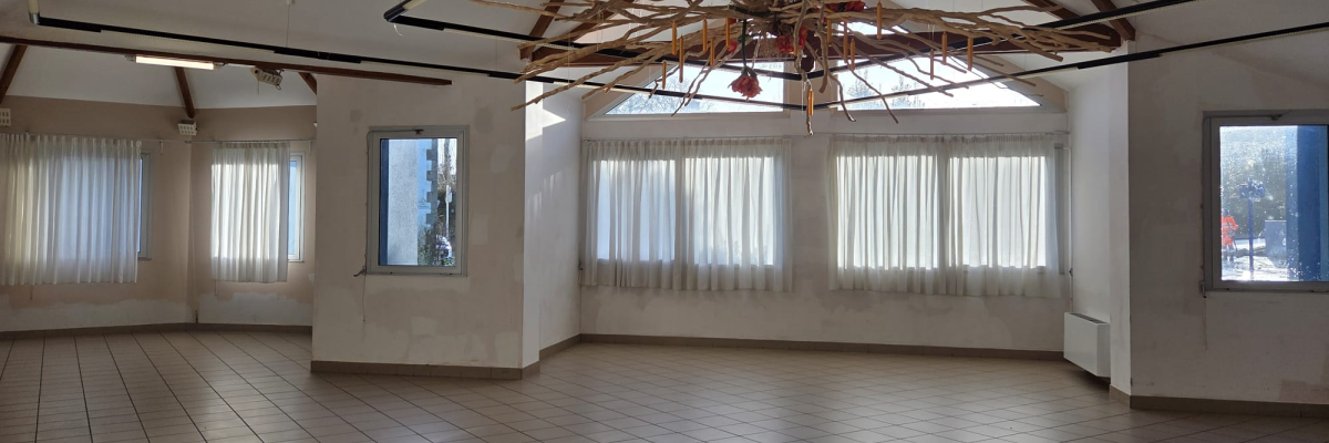 Salle vide avec sol carrelé, fenêtres, lustre et poutres apparentes.