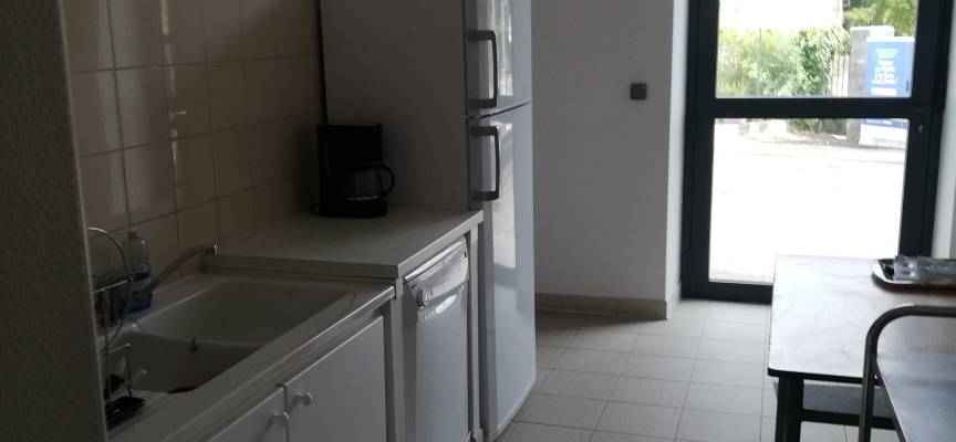 Cuisine avec évier, réfrigérateur, four, micro-ondes, table et chaises. Porte vitrée menant à l'extérieur. Carrelage au sol.