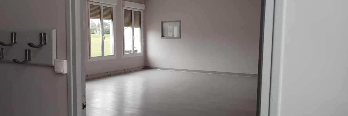 Pièce vide avec parquet, fenêtres, plinthes électriques et tableau.