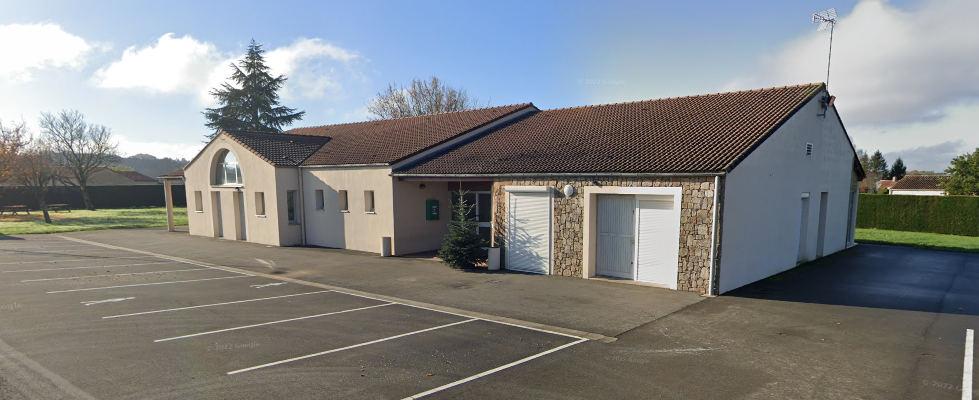 Bâtiment avec toiture en tuiles, façade mixte pierre et enduit, porte de garage blanche, parking marqué, arbres.
