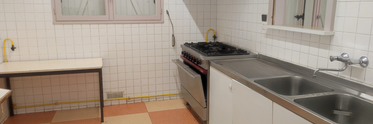 Cuisine avec gaziniere, hotte, évier, placard, fenêtre et carrelage.