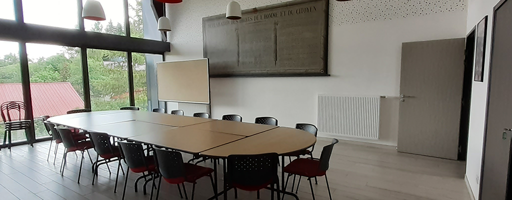 Salle avec table, chaises, tableau blanc, radiateur, porte et suspensions.