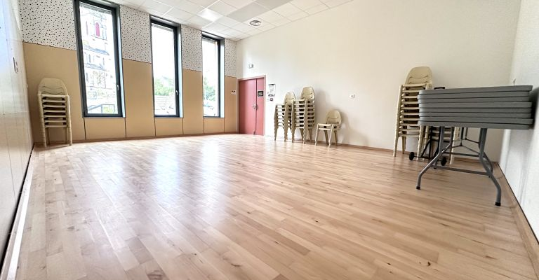 Salle avec parquet, chaises pliantes, tables empilées, miroir et fenêtres.