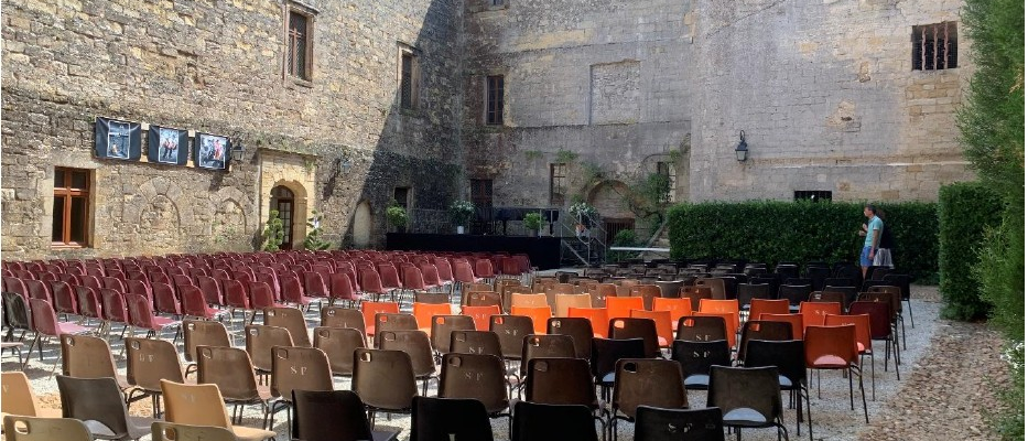 Cour de château avec chaises disposées pour un événement, fond sonore, murs de pierre, fenêtres et plantes.