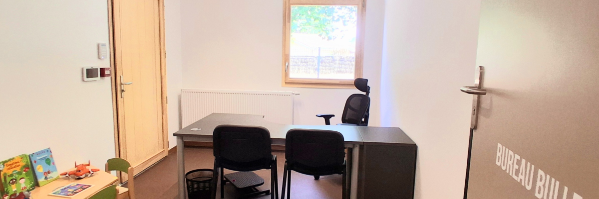 Bureau avec table, chaises, radiateur, fenêtre, panier de basket miniature, petite table avec des jouets, porte avec poignée et plaque "BUREAU BULLE".