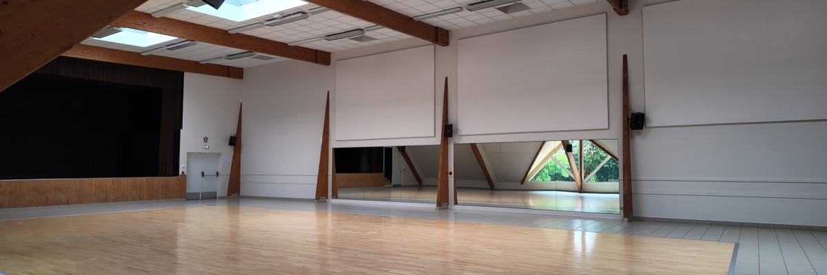 Salle avec sol en bois, murs blancs, poutrelles apparentes, écran, fenêtres et éclairage naturel.