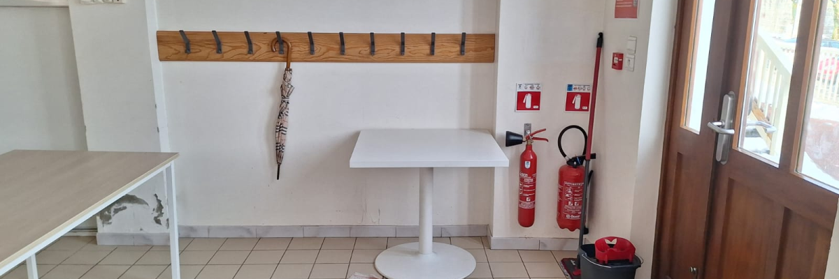 Pièce avec table haute, banc, extincteurs, seau, porte et crochets au mur.