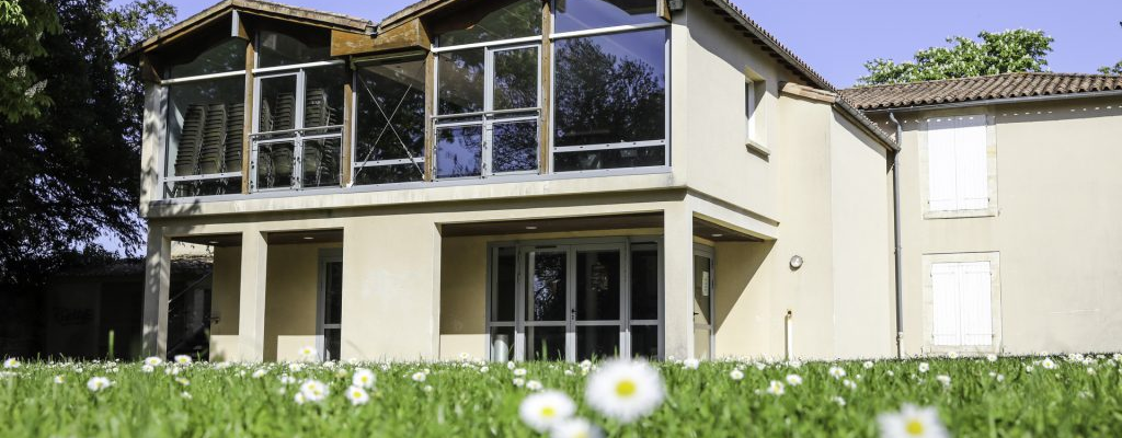 Maison avec étage, balcon et grandes fenêtres. Pelouse avec fleurs blanches.