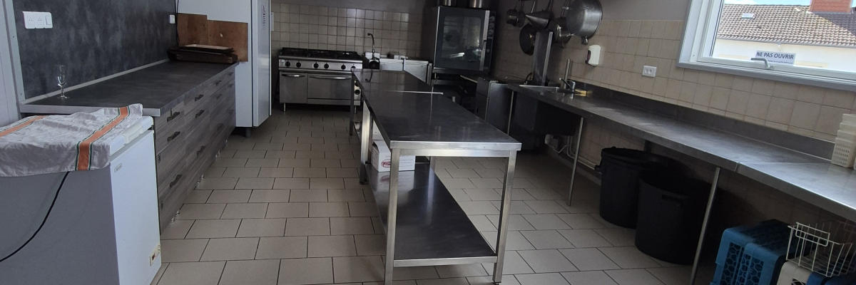 Cuisine industrielle avec plans de travail, éviers, frigo, fours, et poubelles.