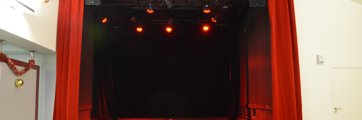 Scène avec rideaux rouges, projecteurs et boule à miroir. Chaises alignées devant.