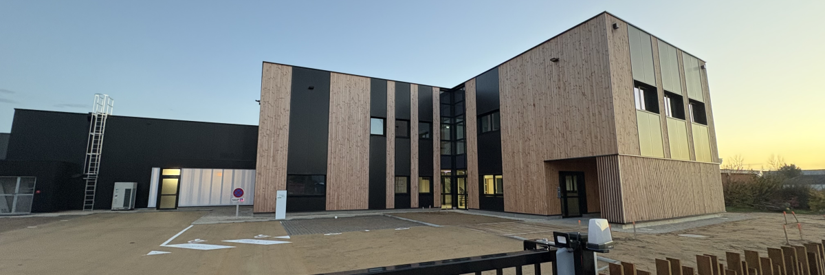 Bâtiment avec façade en bois et panneaux noirs, parking vide, clôture.