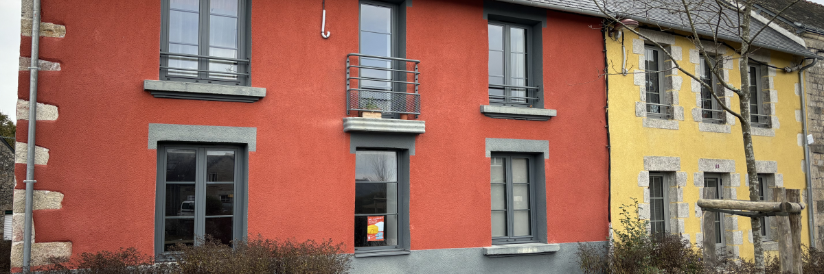 Maison rouge à deux étages avec balcons, vélo devant, arbustes et toits en ardoise.