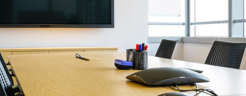 Salle de réunion avec table, chaises, écran, souris, clavier, stylo, lunettes, et rideaux.