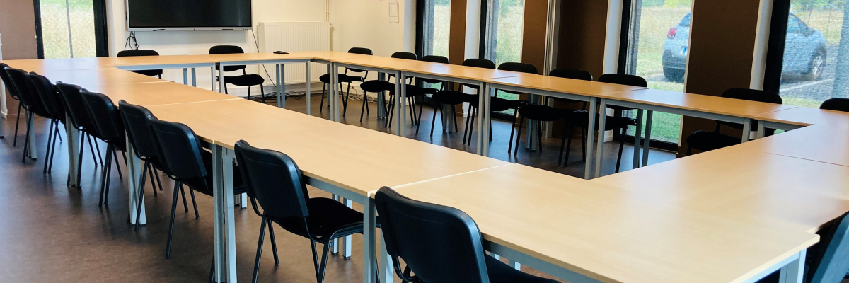 Salle de réunion avec tables en U, chaises, écran et fenêtres.