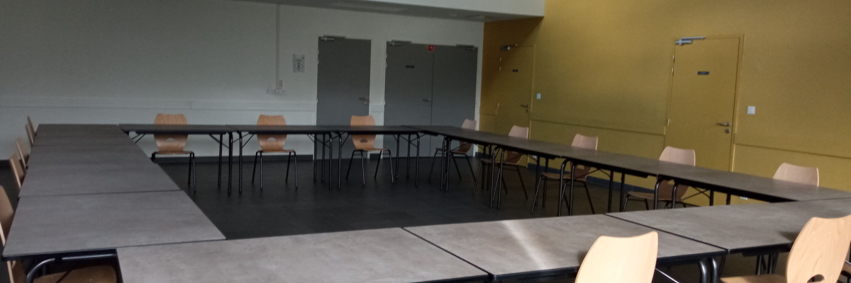 Salle avec tables disposées en U, chaises, murs gris et jaune, portes, et éclairage au plafond.