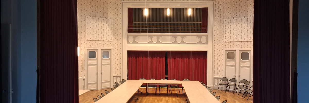 Salle avec scène, rideaux rouges, tables en U, chaises, porte et lumières suspendues.